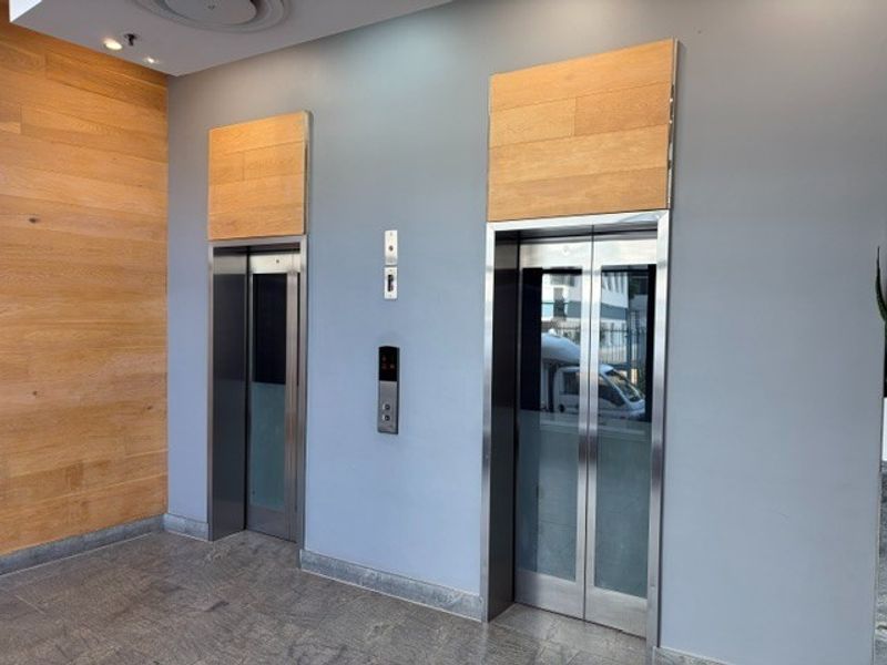 Modern office building lobby featuring dual elevators with stainless steel doors, wood paneling accents, and polished stone flooring.