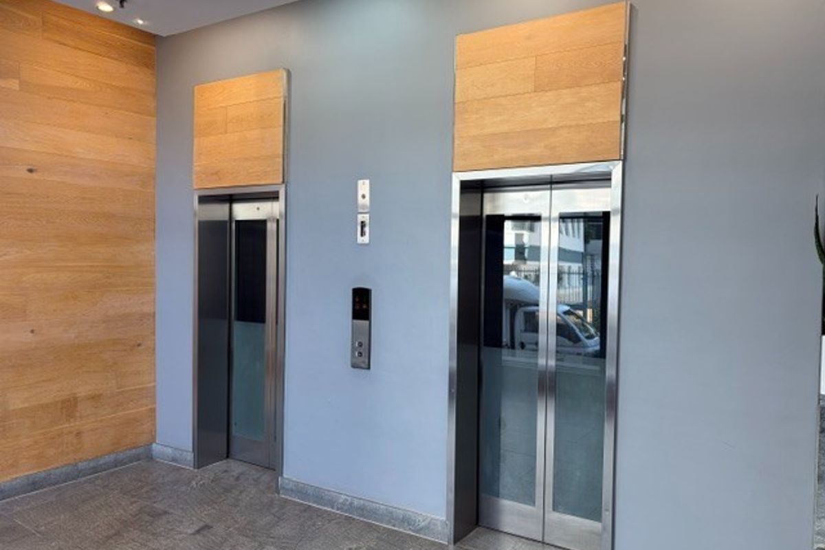 Modern office building lobby featuring dual elevators with stainless steel doors, wood paneling accents, and polished stone flooring.