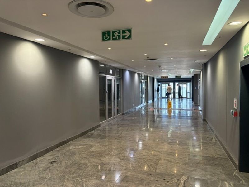 A spacious, well-lit office building hallway featuring polished marble flooring, recessed ceiling lighting, and glass partition doors leading to office suites.