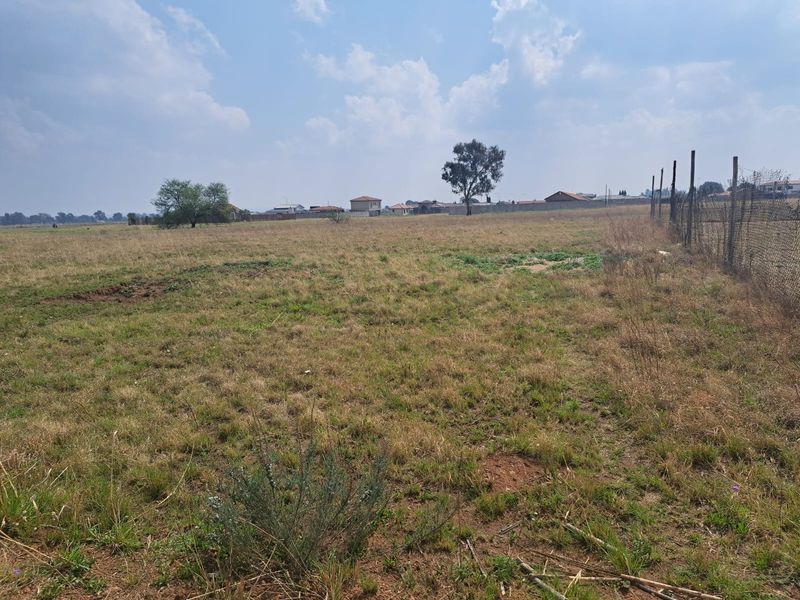 A wide, open vacant residential plot featuring natural grass coverage and perimeter wire mesh fencing, suitable for future development.