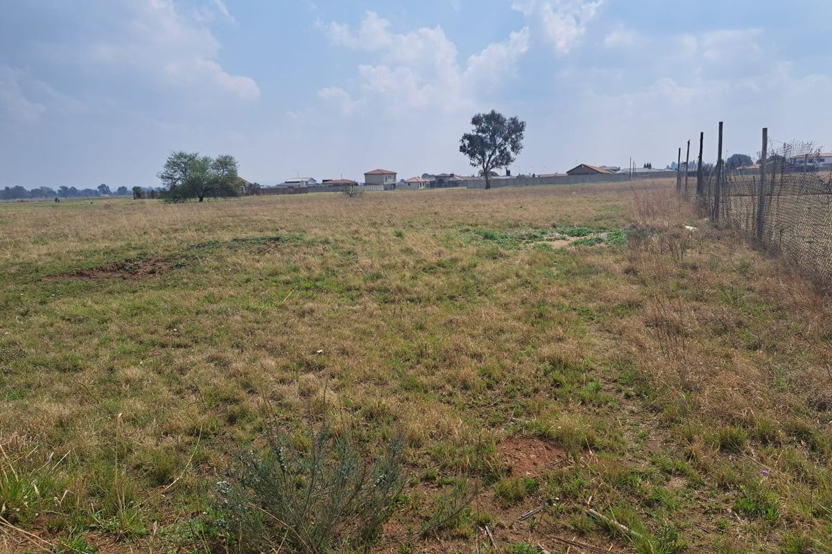 A wide, open vacant residential plot featuring natural grass coverage and perimeter wire mesh fencing, suitable for future development.