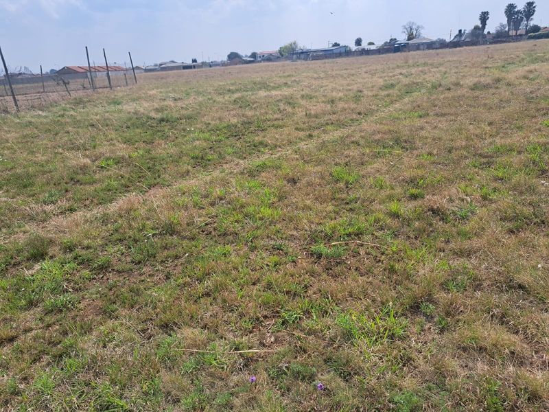 A wide, open vacant residential plot featuring natural grass coverage and perimeter wire mesh fencing in a suburban setting.