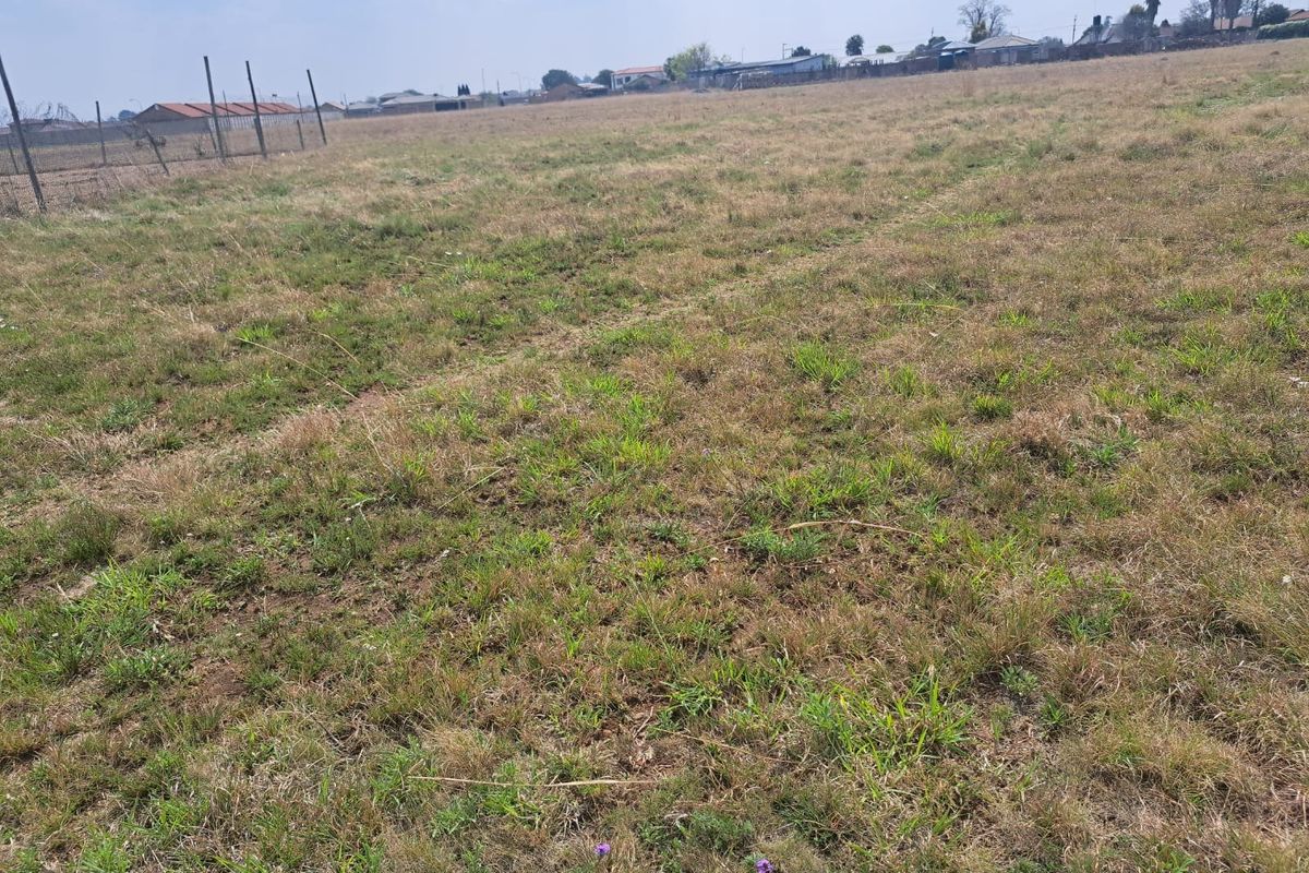 A wide, open vacant residential plot featuring natural grass coverage and perimeter wire mesh fencing in a suburban setting.