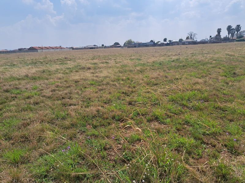 A wide-angle view of a vacant residential plot featuring open grassland with surrounding neighborhood structures in the distance.