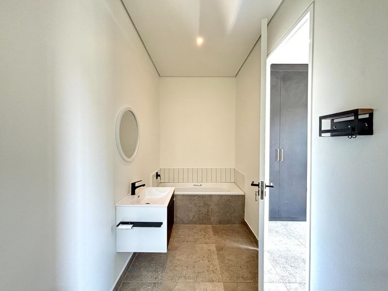 A modern, minimalist bathroom featuring a wall-mounted vanity, a built-in bathtub with tiled surround, and neutral floor tiles.