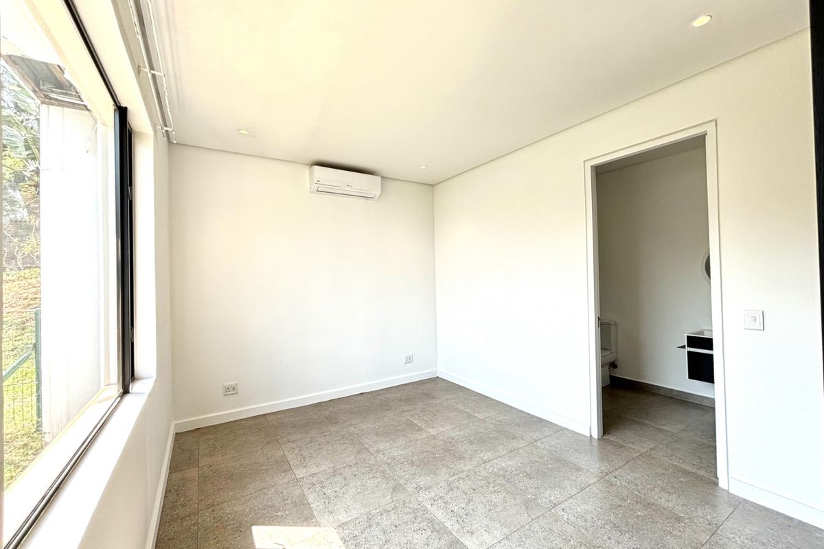A bright, empty bedroom featuring neutral tiled flooring, recessed lighting, and a wall-mounted air conditioning unit, with direct access to an en-suite bathroom.