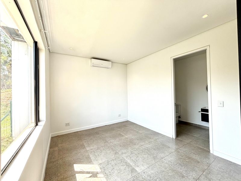 A bright, empty bedroom featuring neutral tiled flooring, recessed lighting, and a wall-mounted air conditioning unit, with direct access to an en-suite bathroom.