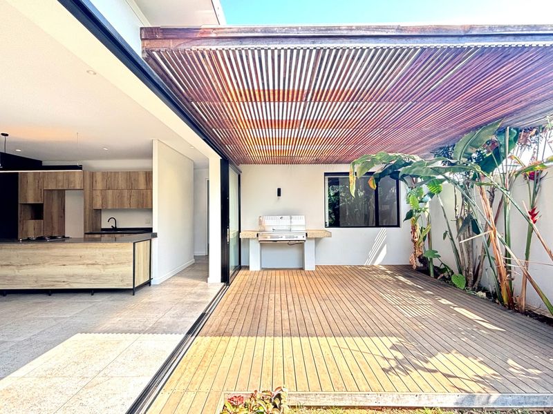 A spacious outdoor patio featuring timber decking, a slatted timber pergola, and a built-in braai area, seamlessly connecting to the open-plan kitchen interior.