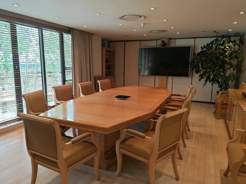 A professional boardroom featuring hardwood flooring, recessed lighting, large windows with blinds, and a wall-mounted display screen for presentations.