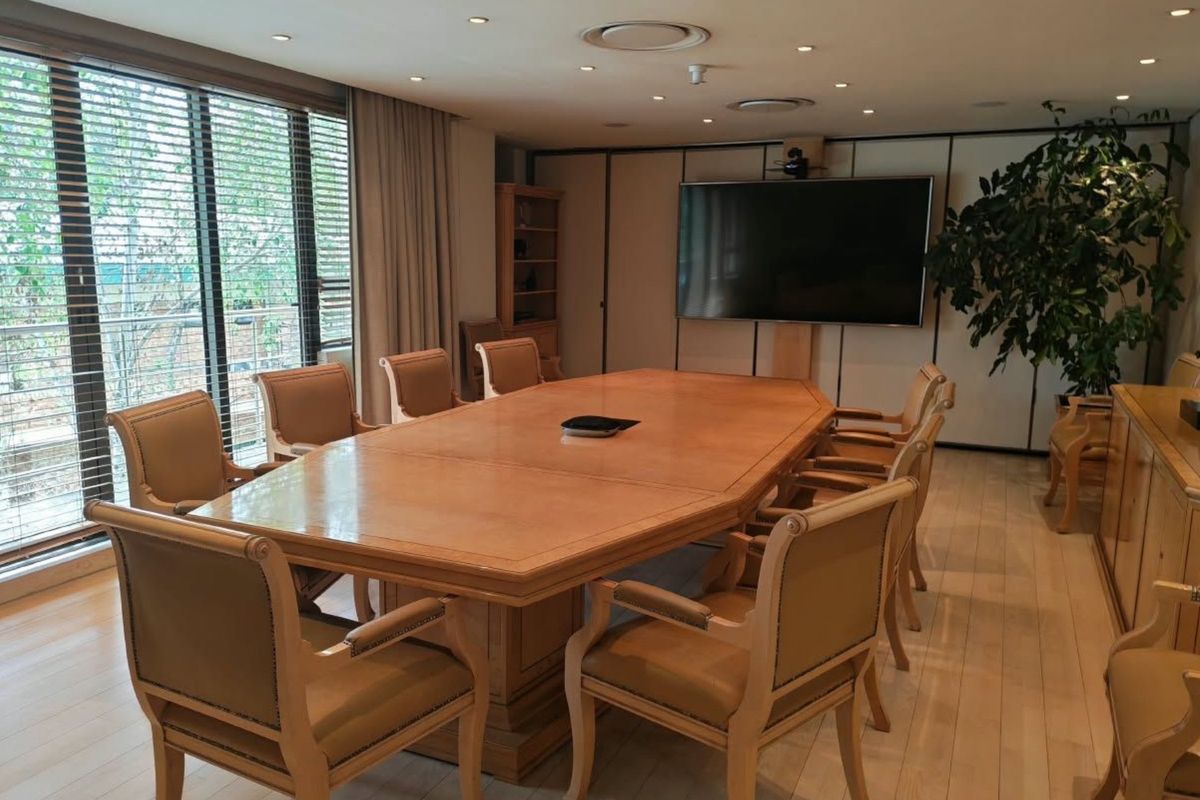 A professional boardroom featuring hardwood flooring, recessed lighting, large windows with blinds, and a wall-mounted display screen for presentations.