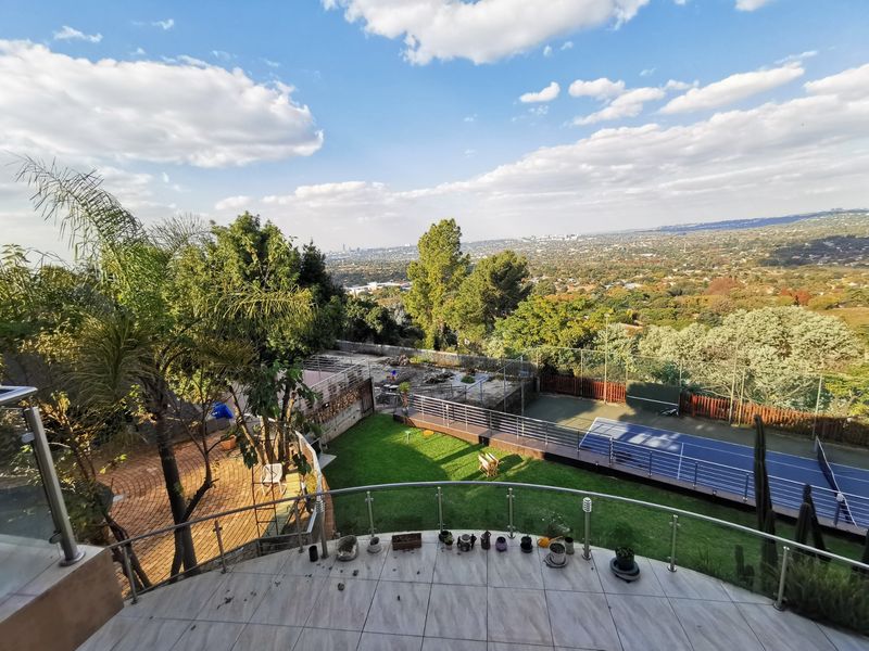 Elevated view overlooking a private tennis court, landscaped garden, and expansive suburban landscape from a tiled balcony with a modern glass balustrade.