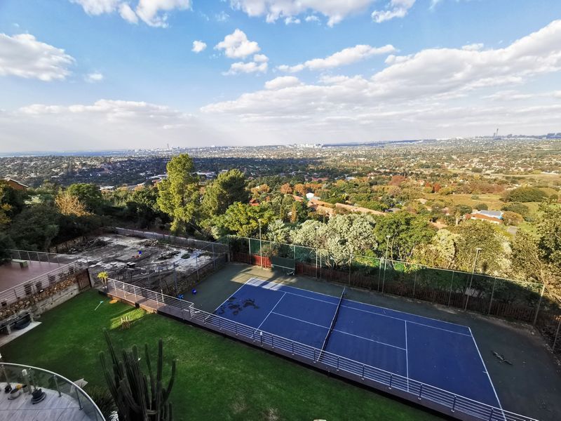 Elevated view of a private tennis court with perimeter fencing, set within a landscaped garden overlooking an expansive urban landscape.