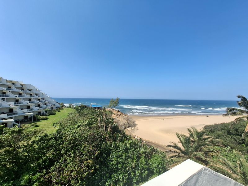 A scenic elevated view overlooking the coastline, sandy beach, and ocean, featuring a clear perspective of the surrounding apartment complex and natural landscape.