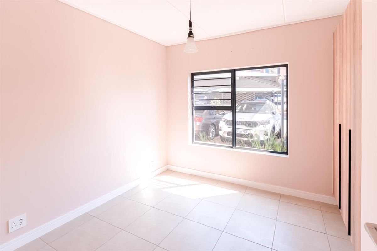 A bright, empty bedroom featuring neutral tiled flooring, soft pink walls, a built-in wardrobe, and an aluminium-framed window with security bars.