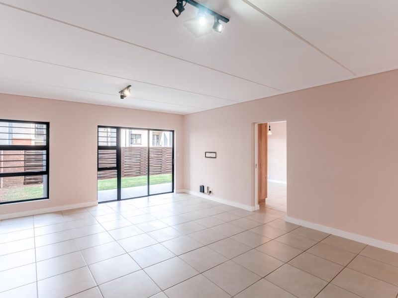 A bright, empty living room featuring neutral tiled flooring, modern track lighting, and large sliding glass doors providing access to the outdoor area.