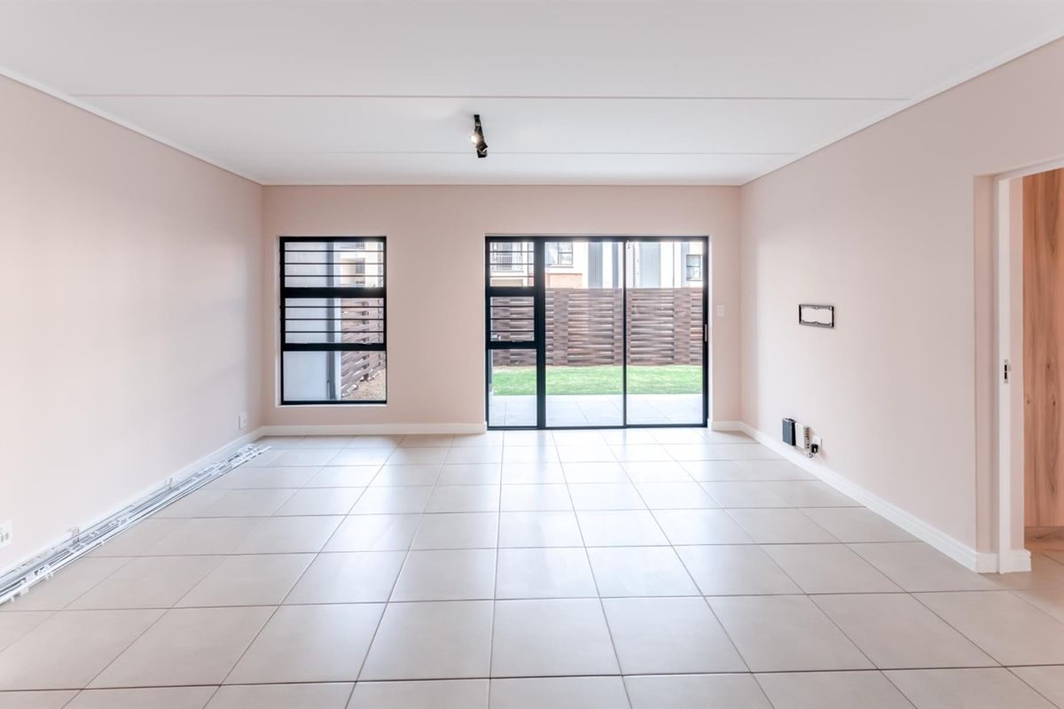 A bright, empty living room featuring neutral tiled flooring, modern track lighting, and a sliding glass door providing access to the rear garden area.