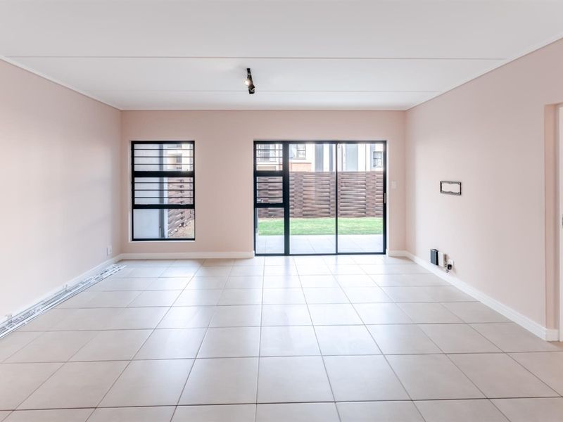 A bright, empty living room featuring neutral tiled flooring, modern track lighting, and a sliding glass door providing access to the rear garden area.