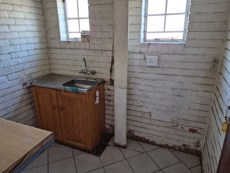 A functional scullery area featuring a stainless steel sink, wooden cabinetry, tiled flooring, and exposed brick walls in need of cosmetic refurbishment.