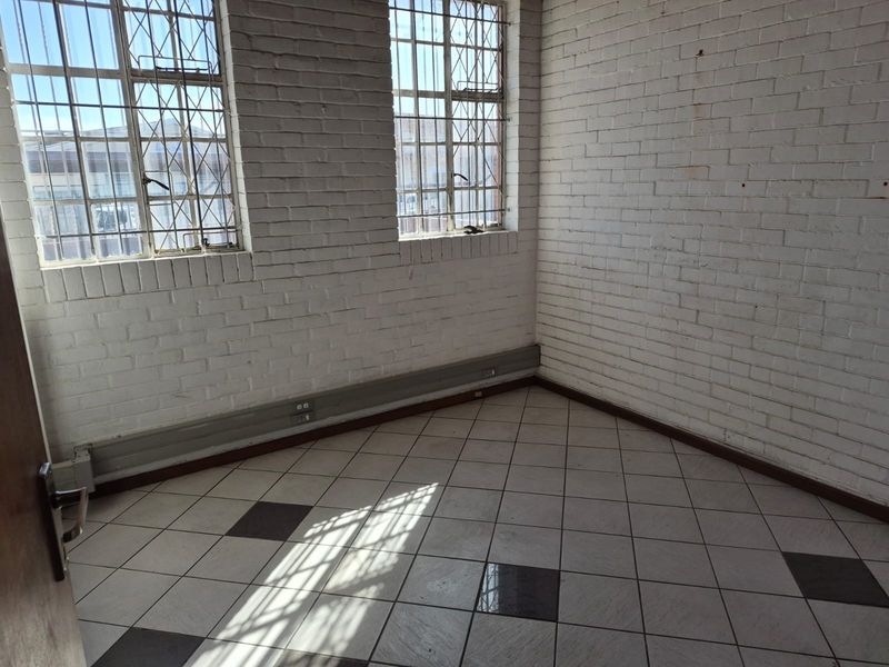 An empty industrial office space featuring exposed white brick walls, tiled flooring, and windows fitted with security bars.