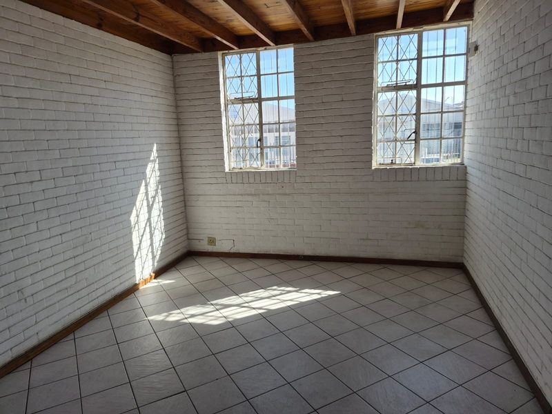 An empty office space featuring exposed timber ceiling beams, painted brick walls, tiled flooring, and windows fitted with security bars.