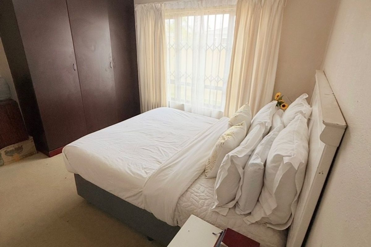 A functional bedroom featuring built-in dark wood cupboards, neutral wall finishes, and a window fitted with security bars.