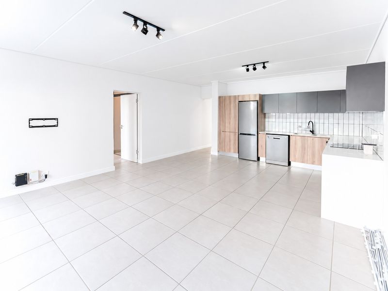A bright, modern open-plan living and kitchen area featuring neutral tiled flooring, contemporary track lighting, and sleek wood-tone and grey cabinetry.