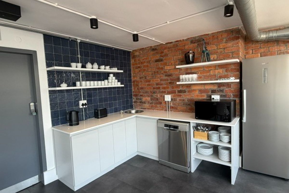 Modern office kitchenette featuring exposed brickwork, blue tiled splashback, white cabinetry, integrated dishwasher, and industrial-style exposed ducting and track lighting.