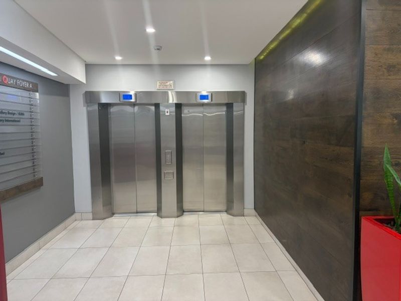 Modern office building lobby featuring dual stainless steel elevators, tiled flooring, and a decorative wood-paneled feature wall.