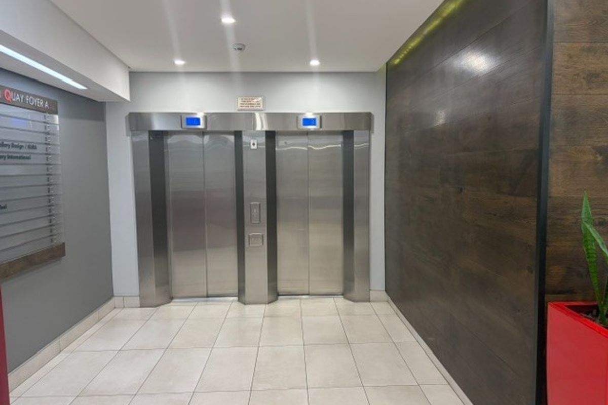 Modern office building lobby featuring dual stainless steel elevators, tiled flooring, and a decorative wood-paneled feature wall.
