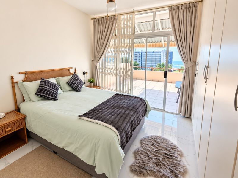 A bright bedroom featuring built-in wardrobes, tiled flooring, and a sliding glass door providing direct access to a private balcony with ocean views.