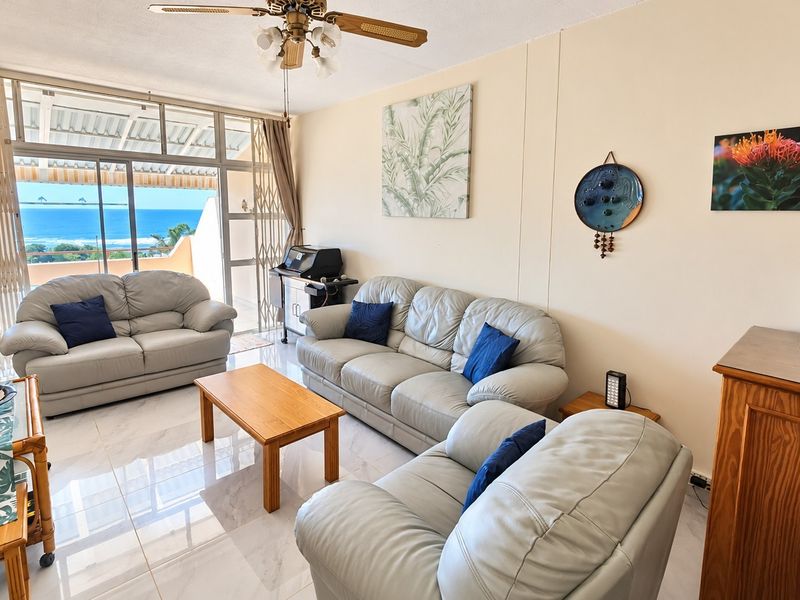 A bright living room featuring tiled floors, a ceiling fan, and large sliding glass doors that open to a balcony with ocean views.