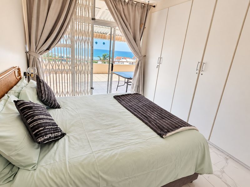 A bright bedroom featuring built-in cupboards, tiled flooring, and a sliding glass door leading to a balcony with ocean views.