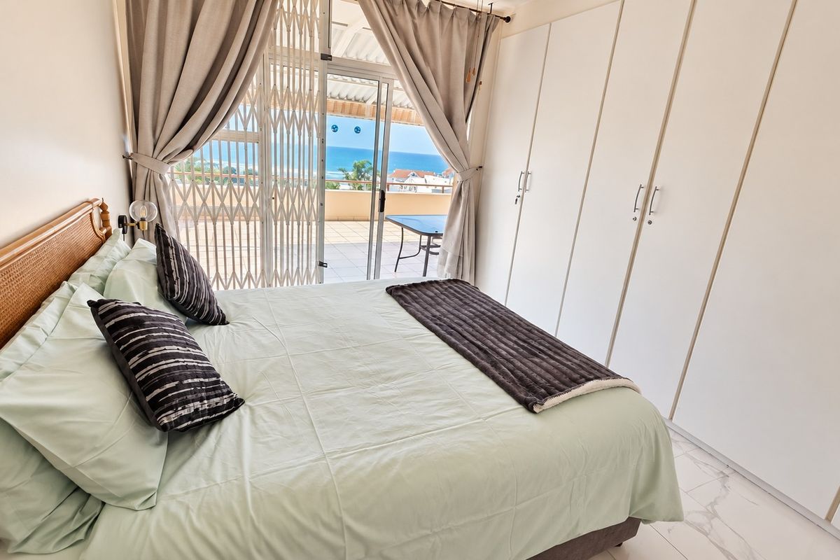 A bright bedroom featuring built-in cupboards, tiled flooring, and a sliding glass door leading to a balcony with ocean views.