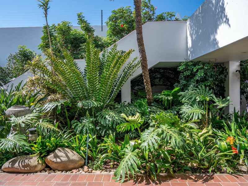 A lush, well-maintained garden area featuring mature tropical plants, brick paving, and a modern white structural staircase.