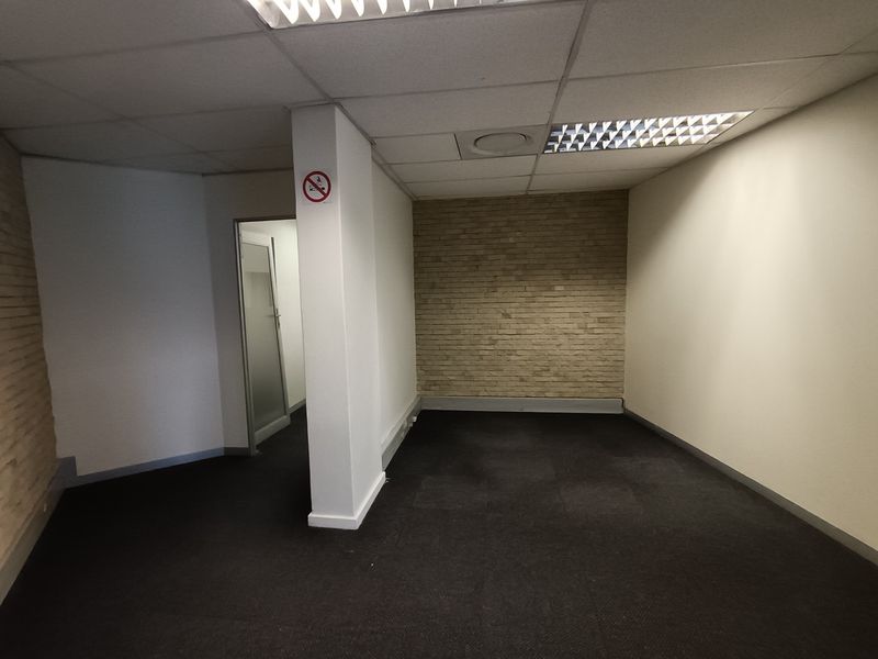 An empty office space featuring a suspended ceiling with recessed lighting, dark carpeting, and a decorative exposed brick feature wall.