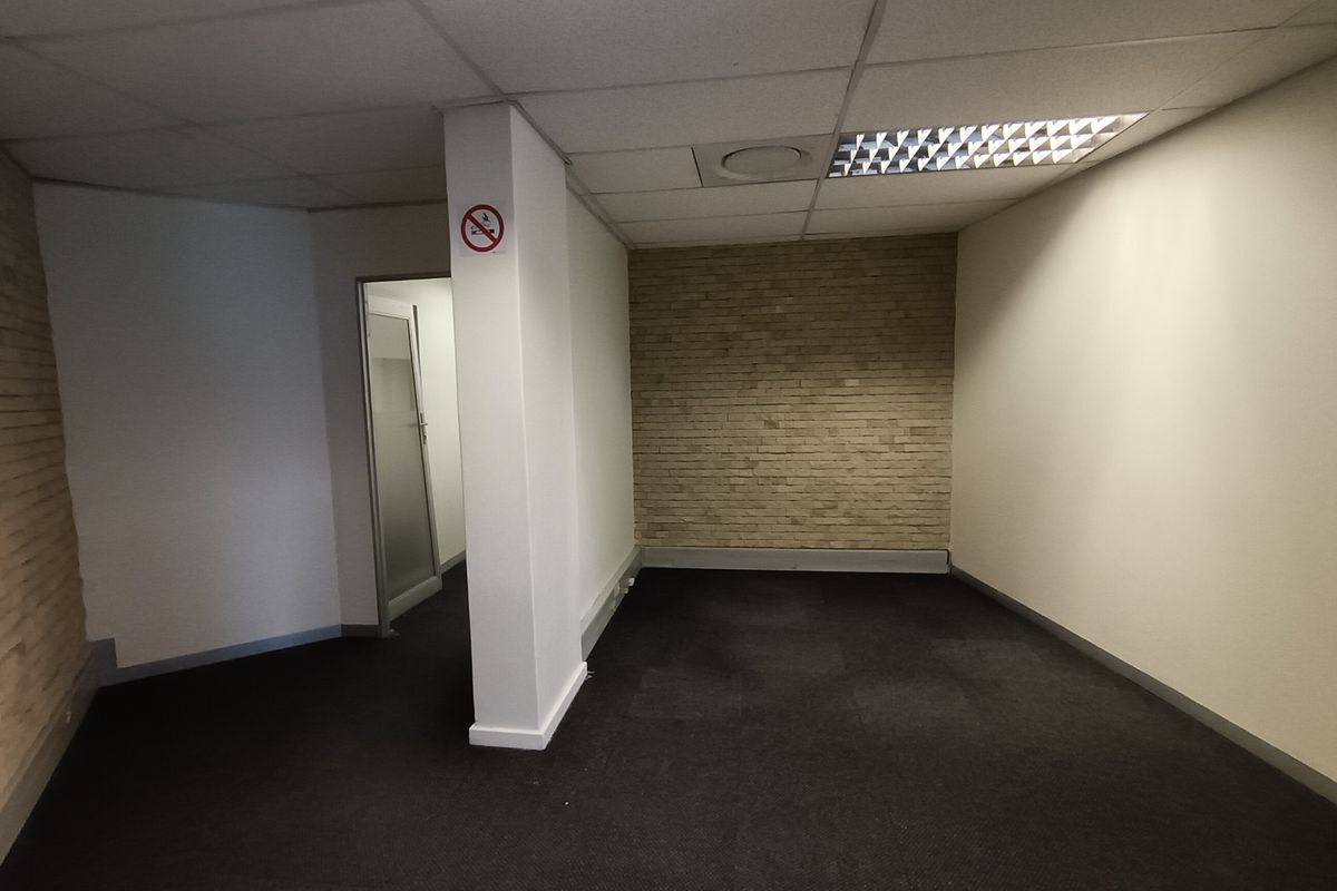 An empty office space featuring a suspended ceiling with recessed lighting, dark carpeting, and a decorative exposed brick feature wall.