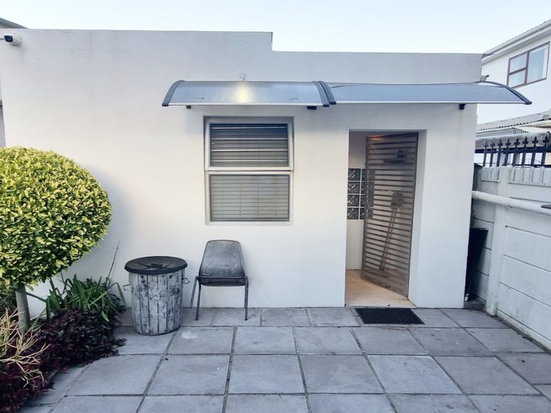 A rear exterior view of the cottage featuring a paved patio area, a protective awning over the entrance, and a secure boundary wall.