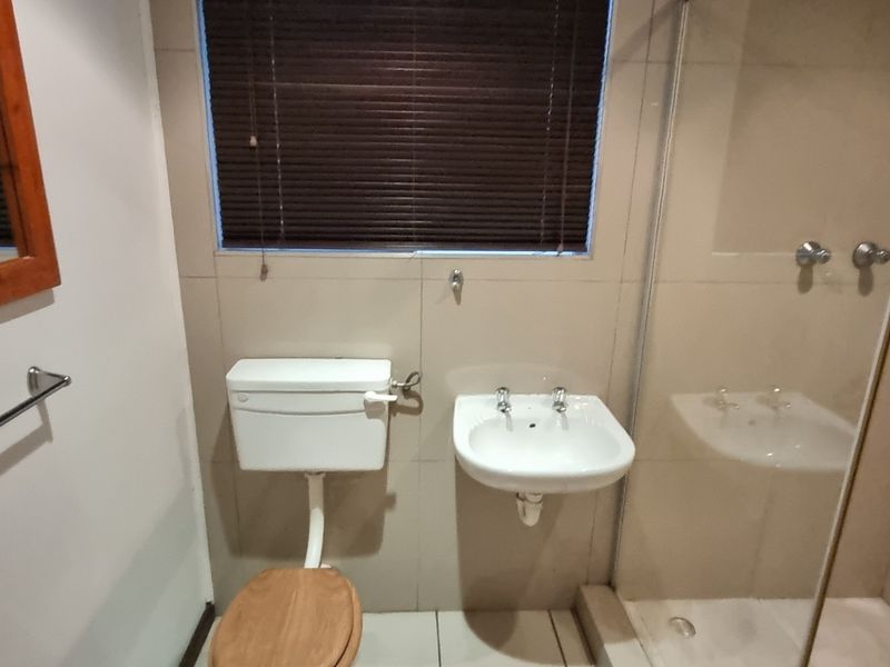 A functional bathroom featuring a wall-mounted basin, toilet, and a glass-enclosed shower with neutral wall tiling and a window with a venetian blind.