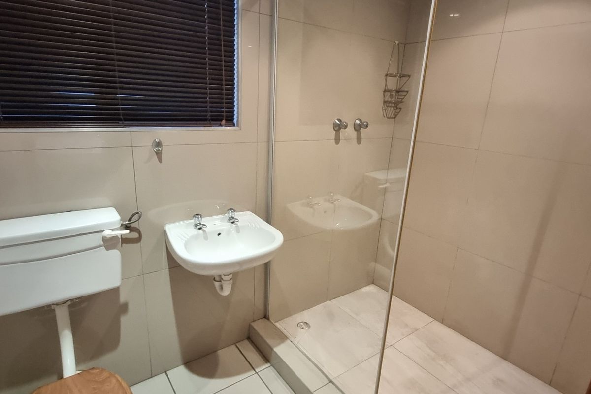 A clean, functional bathroom featuring a wall-mounted basin, toilet, and a glass-enclosed shower with neutral wall and floor tiling.