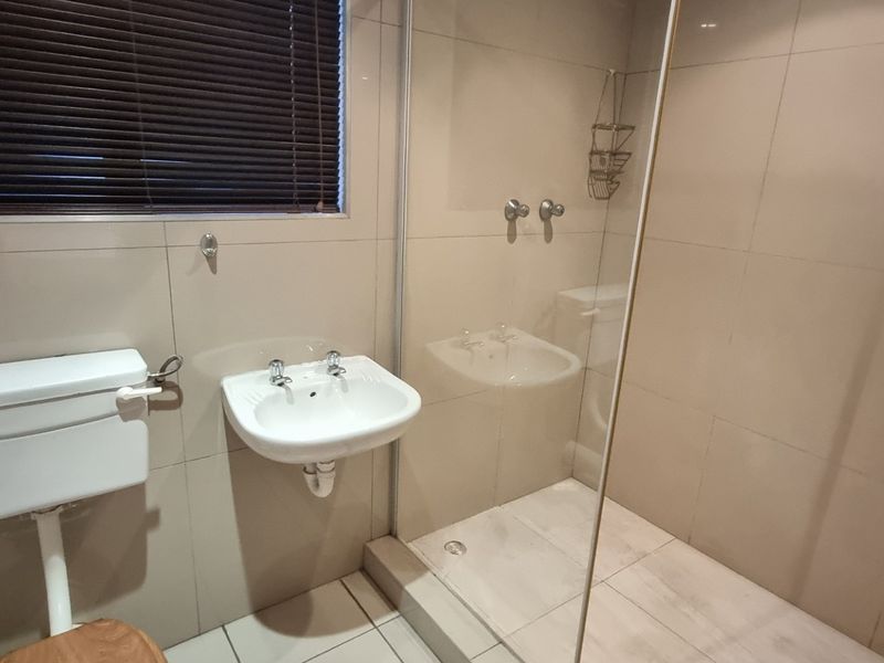 A clean, functional bathroom featuring a wall-mounted basin, toilet, and a glass-enclosed shower with neutral wall and floor tiling.