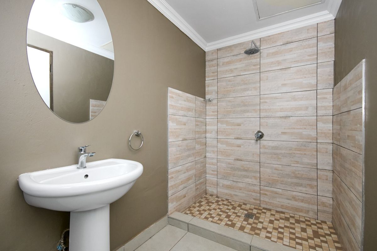 A clean, functional bathroom featuring a pedestal washbasin, an oval mirror, and a walk-in shower with neutral-toned wall and floor tiling.