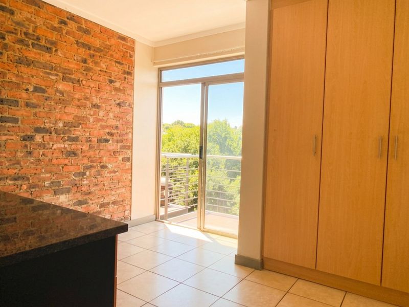 A bright bedroom featuring an exposed brick feature wall, built-in wooden cupboards, tiled flooring, and a sliding door leading to a balcony.