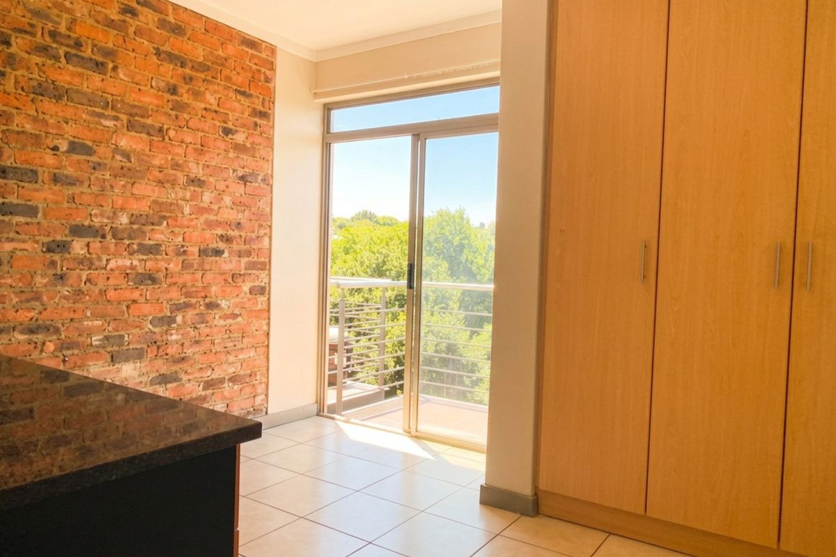 A bright bedroom featuring an exposed brick feature wall, built-in wooden cupboards, tiled flooring, and a sliding door leading to a balcony.