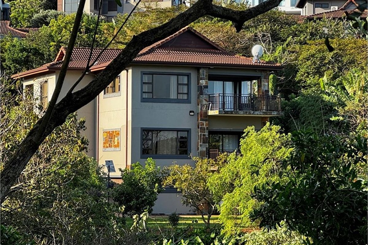 A multi-story residential property featuring a tiled roof, stone cladding accents, and a private balcony, nestled within a lush, mature garden setting.