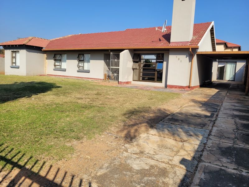 Front view of a single-story house featuring a tiled roof, chimney, paved driveway, and carport, set on a residential lawn.