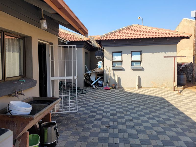A paved rear courtyard area featuring a concrete utility sink, security gate, and exterior wall access, showing signs of wear and maintenance needs.