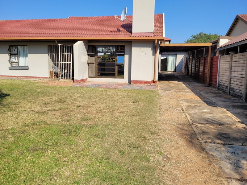 Front view of a single-story house featuring a tiled roof, chimney, paved driveway, and an attached carport with a surrounding lawn.