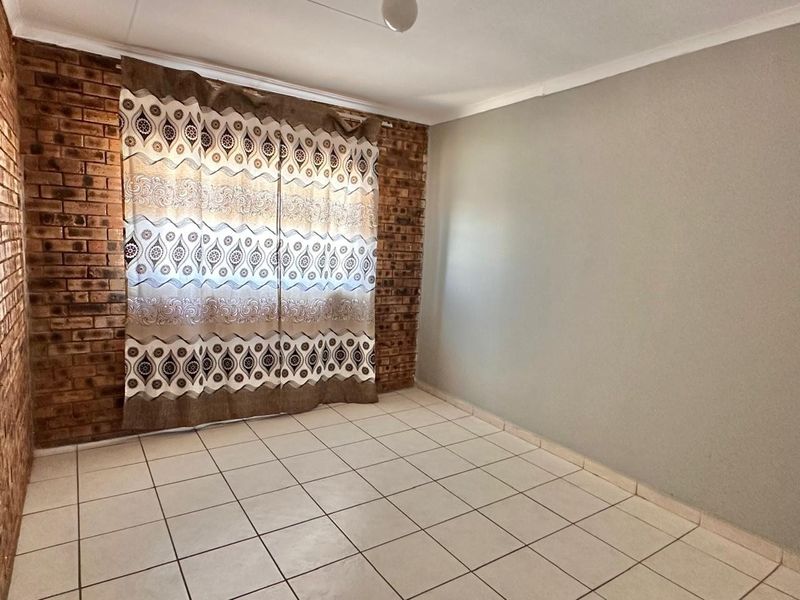 A bright, empty bedroom featuring an exposed brick feature wall, neutral painted walls, and durable ceramic floor tiles.
