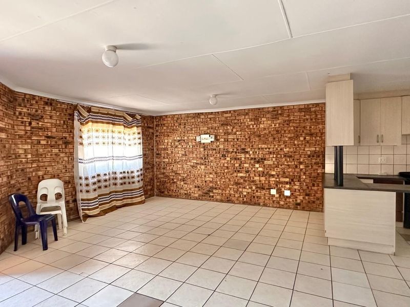 Open-plan living and kitchen area featuring exposed brick walls, ceramic floor tiling, and functional kitchen cabinetry with a laminate countertop.