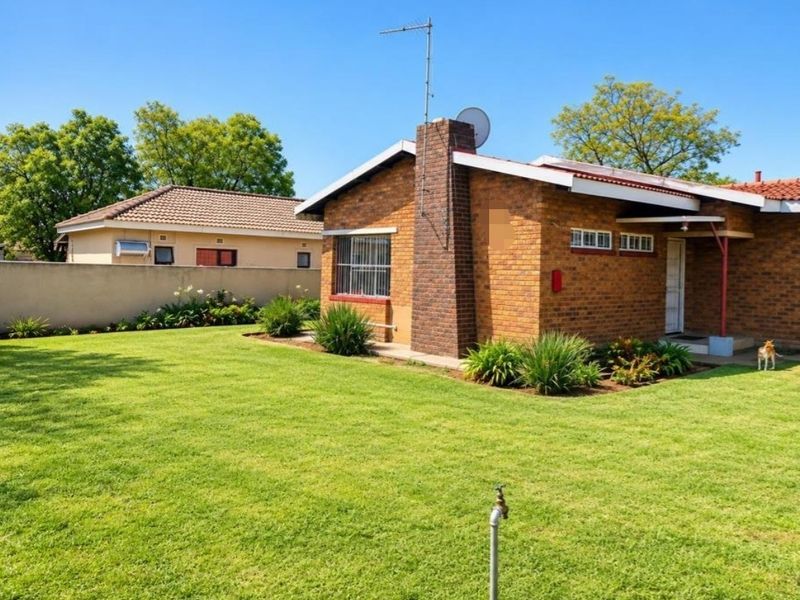 A view of the rear exterior of a brick house featuring a chimney, tiled roof, and a well-maintained lawn with a boundary wall.